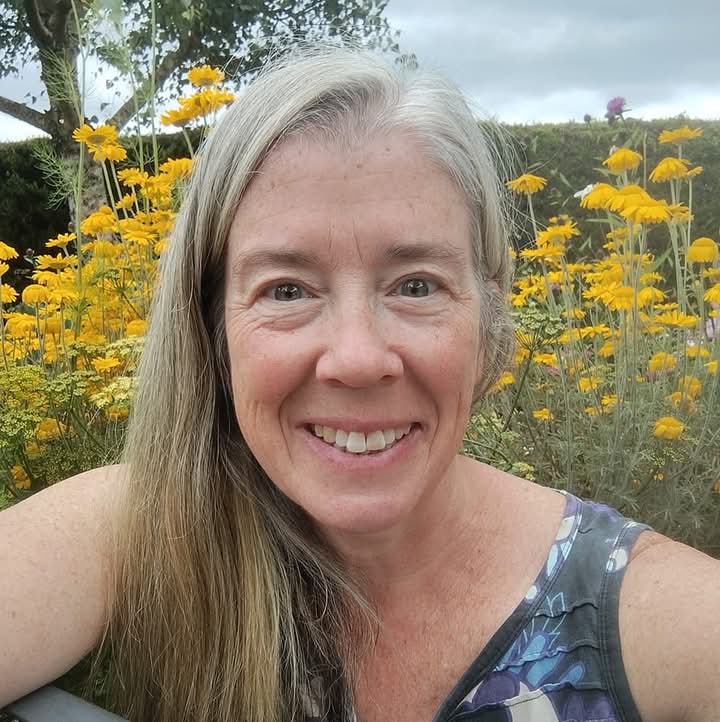 a photo of me - a woman in her 50s with long, grey hair, in the garden, with yellow flowers behind me