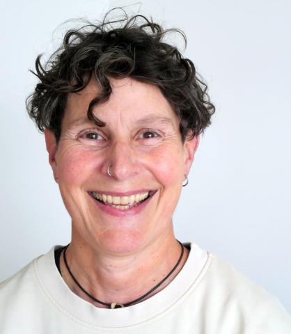 Portrait of a caucasian person in their fifties, with short, dark curly hair and dark eyes. They are smiling into the camera
