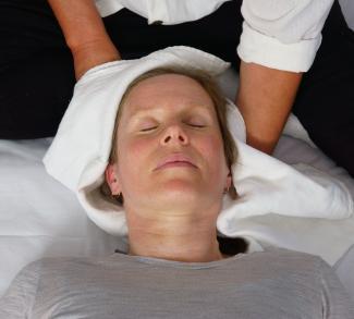 Photo of a caucasian female lying face up on a white cover, eyes closed in a relaxed manner, a pair of hands wrapping a white muslin cloth around her head