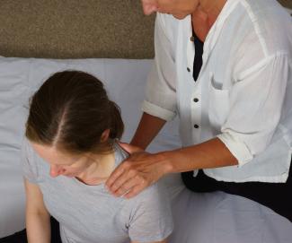 Photo of a caucasian female sitting on a white cover with another person kneeling behind her, one hand on her left shoulder, the other one on the middle of her back