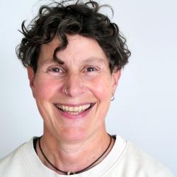 Portrait of a caucasian person in their fifties, with short, dark curly hair and dark eyes. They are smiling into the camera