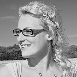 Picture of Louise Shannon shown in black and white, wearing glasses, with a plait in her hair, and a flower crown on her head. She is looking away from the camera. 